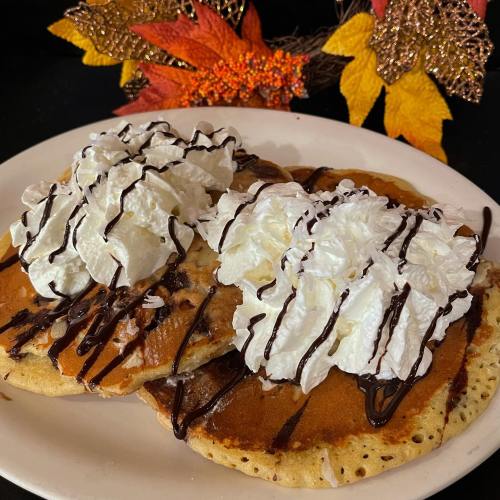 Two fluffy pancakes topped with whipped cream and chocolate drizzle, surrounded by autumn leaves.