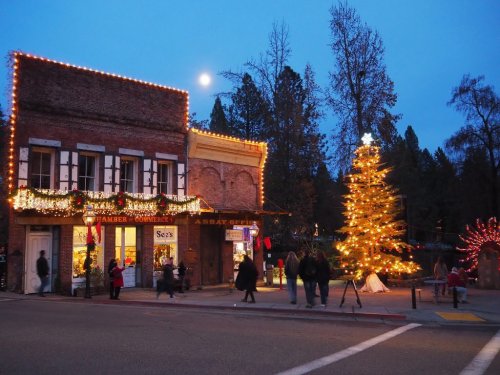 A festive street scene at dusk, featuring a decorated tree and buildings adorned with holiday lights.