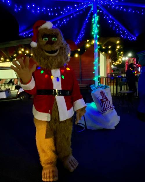 A festive character in a Santa suit with lights, waving under colorful holiday lights.