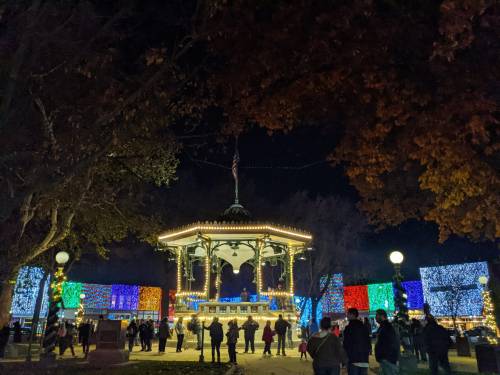 A festive park at night, featuring a decorated gazebo and colorful lights illuminating the surrounding trees and structures.