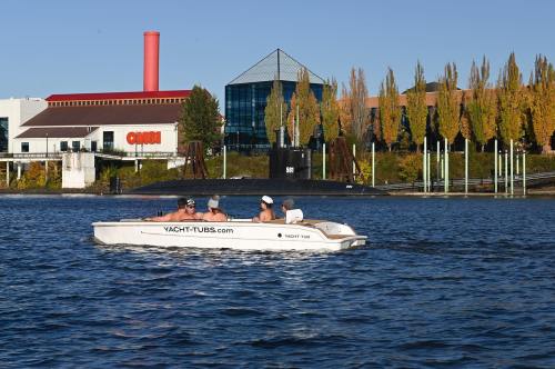 A small boat with four people floats on a lake, with buildings and autumn trees in the background.