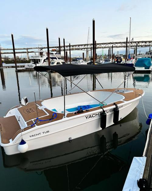A white boat named "Yacht Tub" docked in a marina, reflecting in calm water with other boats in the background.