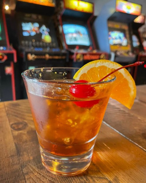 A glass of cocktail with ice, a cherry, and an orange slice, set against a backdrop of vintage arcade machines.