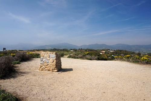 A stone monument stands on a gravel path, surrounded by greenery and mountains under a clear blue sky.