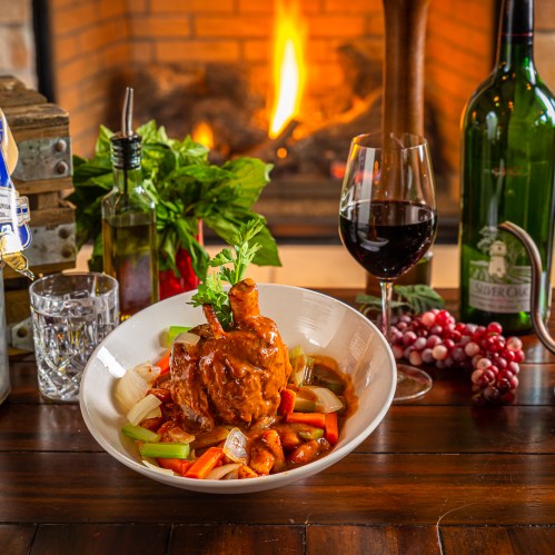 A bowl of savory chicken dish with vegetables, a glass of red wine, and a cozy fireplace in the background.