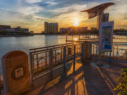 Sunset over a waterfront with buildings in the background, featuring a walkway and informational sign.