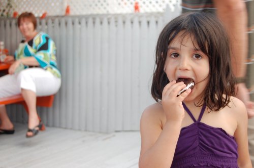 A young girl in a purple top enjoys a treat, while an adult sits in the background.