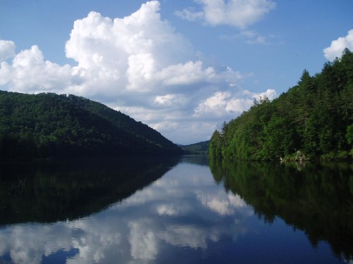 A serene lake surrounded by lush green hills and fluffy clouds reflecting on the water's surface.