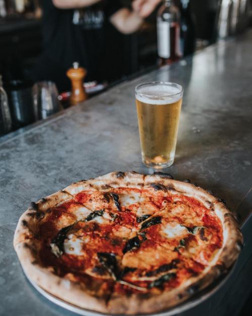 A freshly baked pizza with mozzarella and basil sits on a bar counter next to a glass of beer.