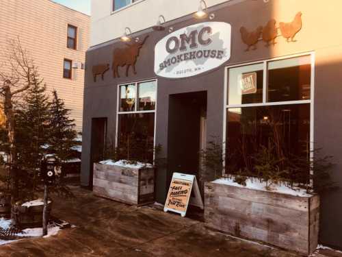 Exterior of OMC Smokehouse in Duluth, MN, featuring a sign and planters with small trees in a snowy setting.