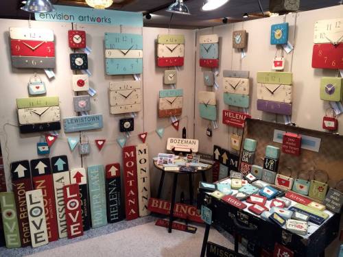 Artisan booth displaying colorful clocks and wooden signs with city names and decorative elements.