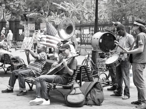 A group of musicians playing instruments in a park, with a backdrop of trees and art displays.