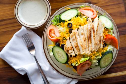 A colorful salad topped with grilled chicken, tomatoes, cucumbers, olives, and shredded cheese on a wooden table.