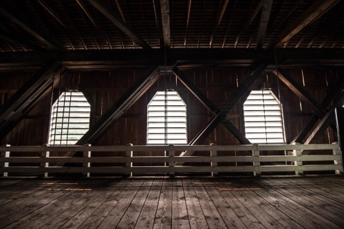 Interior of a rustic barn with wooden beams and slatted windows, casting light onto the wooden floor.