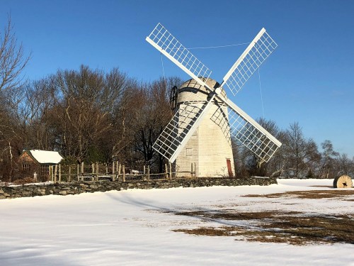 A white windmill with large sails stands beside a snowy field, surrounded by trees under a clear blue sky.