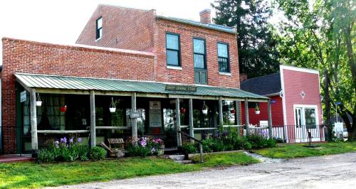 Historic brick building with a green awning, surrounded by flowers and trees, featuring a red building nearby.
