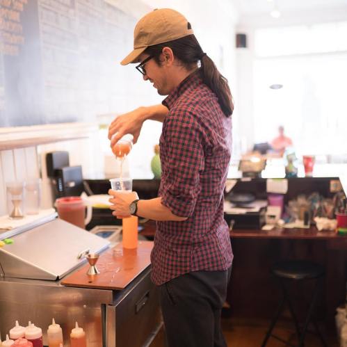 A person in a plaid shirt and cap prepares a drink at a café, with various condiments and equipment in the background.