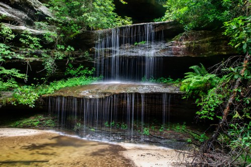 A serene waterfall cascading over rocky ledges, surrounded by lush greenery and a sandy area below.