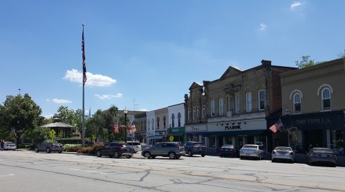 A sunny street view of a small town with shops, parked cars, and American flags lining the buildings.