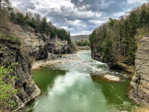 A serene river flows between rocky cliffs, surrounded by lush trees under a cloudy sky.