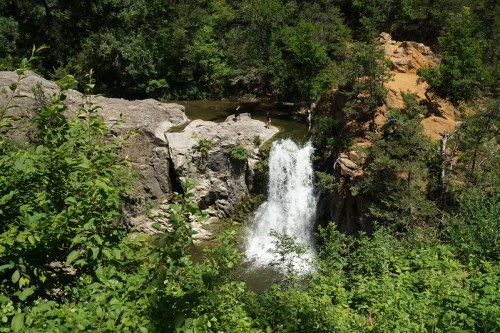 A scenic waterfall cascades over rocks, surrounded by lush greenery and trees in a natural setting.