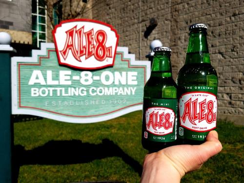 A hand holds two bottles of Ale-8-One soft drink in front of the Ale-8-One Bottling Company sign.
