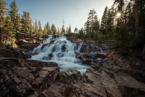A serene waterfall cascades over rocky terrain, surrounded by tall trees and bathed in soft sunlight.
