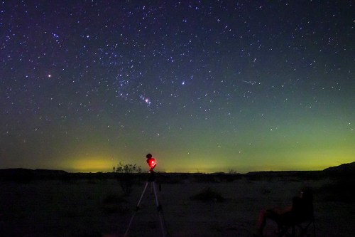 A person sits in a chair under a starry night sky, with a telescope pointed at the stars and a faint green glow on the horizon.