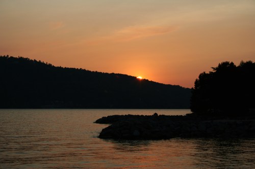 Sunset over a calm lake, with silhouettes of trees and mountains against a colorful sky.
