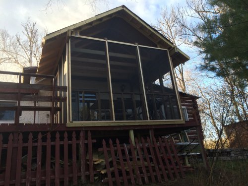 A wooden house with a screened porch, surrounded by trees and a wooden fence, under a partly cloudy sky.