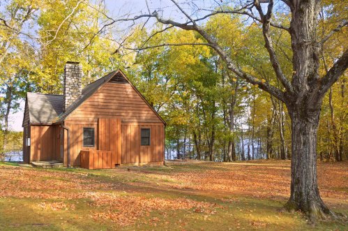 A rustic wooden cabin surrounded by colorful autumn foliage and fallen leaves, near a tranquil lake.