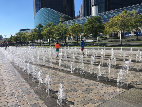 Two children play in a fountain area with jets of water, surrounded by trees and modern buildings on a sunny day.