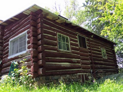 A rustic log cabin with a stone foundation, surrounded by greenery and trees under a clear sky.