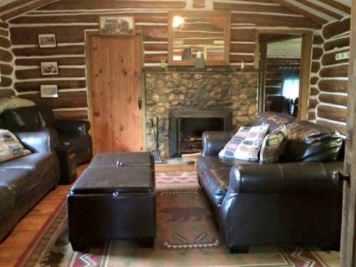 Cozy log cabin interior with stone fireplace, leather sofas, and a coffee table on a patterned rug. Warm and inviting atmosphere.