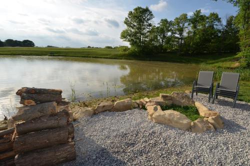 A serene lakeside scene with two chairs, a stone fire pit, and lush greenery in the background.