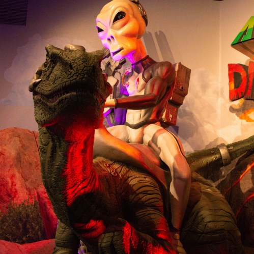A cartoonish alien riding a dinosaur in a colorful, whimsical setting.