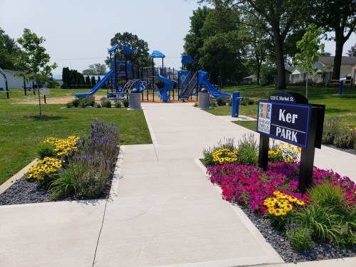 A playground with blue equipment surrounded by colorful flowers and a sign reading "Ker Park." Sunny day.