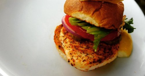 A grilled chicken sandwich with lettuce, tomato, onion, and a creamy sauce on a toasted bun, served on a white plate.