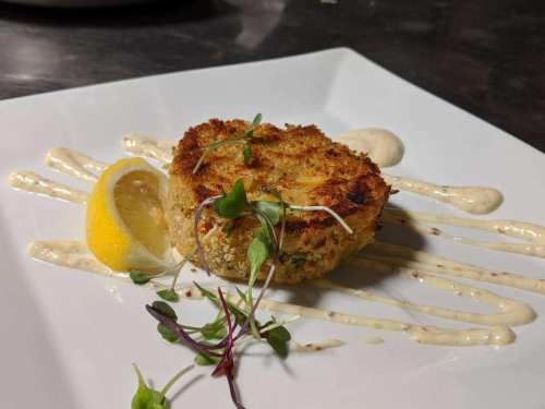 A golden-brown crab cake on a white plate, garnished with microgreens and a lemon wedge, drizzled with sauce.