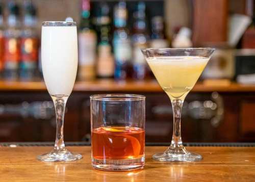 Three cocktails on a bar: a frothy white drink, a yellow martini, and a dark amber drink in a low glass.