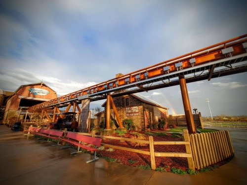 A roller coaster track extends from a rustic building under a cloudy sky, with a rainbow in the background.