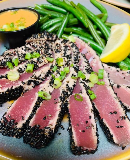 Sliced seared tuna with sesame crust, green beans, a lemon wedge, and a small cup of sauce on a plate.