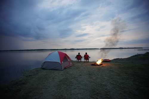 Two people sit by a calm lake near a tent, watching a fire as smoke rises against a cloudy sky.