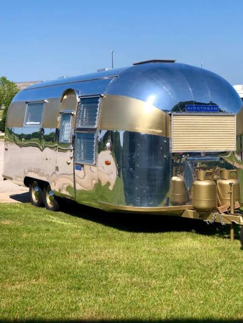 A shiny, vintage Airstream trailer parked on green grass, reflecting its surroundings in the polished surface.