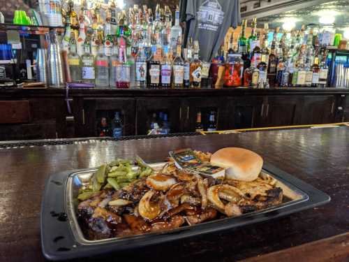 A plate of grilled meat with onions, a bun, and green beans, set on a bar counter with a variety of liquor bottles in the background.