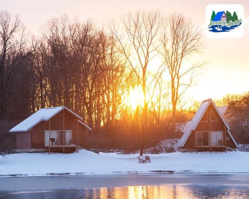 Two cozy cabins by a snowy lake at sunset, surrounded by trees. A serene winter landscape.
