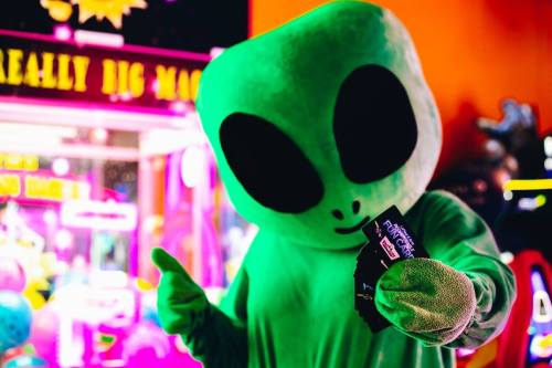 A green alien mascot holding game tickets and giving a thumbs up in a colorful arcade setting.