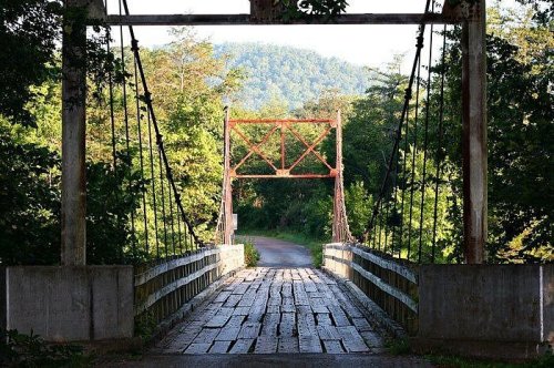 A rustic suspension bridge spans a tree-lined path, surrounded by lush greenery and distant hills.