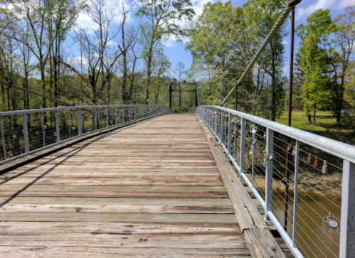 A wooden pedestrian bridge stretches over a calm river, surrounded by lush green trees and a clear blue sky.