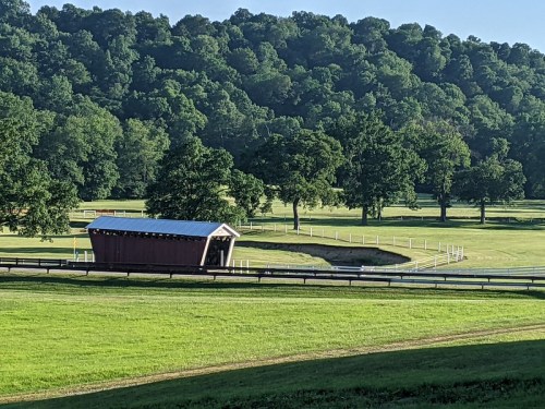 A scenic view of a covered bridge beside a winding stream, surrounded by lush green fields and trees.